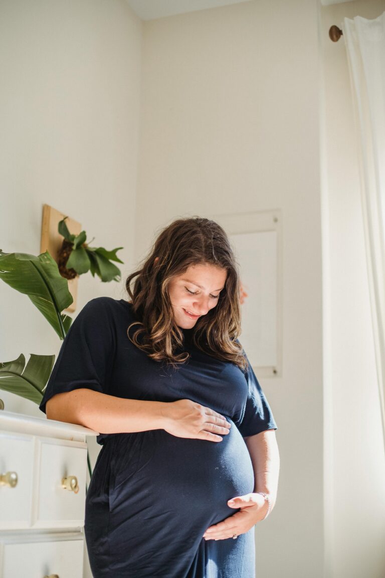 Pregnant woman with brown hair and a blue dress holding belly in her hands, looking at baby bump, and smiling | Unexpected Pregnancy | Mary's Refuge | Martinsburg WV