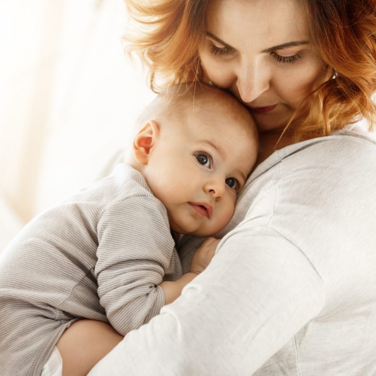 Beautiful young mother tenderly hugs her little cute child. Mom looking at her kid and smiling while baby looks aside and snuggling to mom chest. Happy family moments. | Stories of Hope | Mary's Refuge | Martinsburg WV