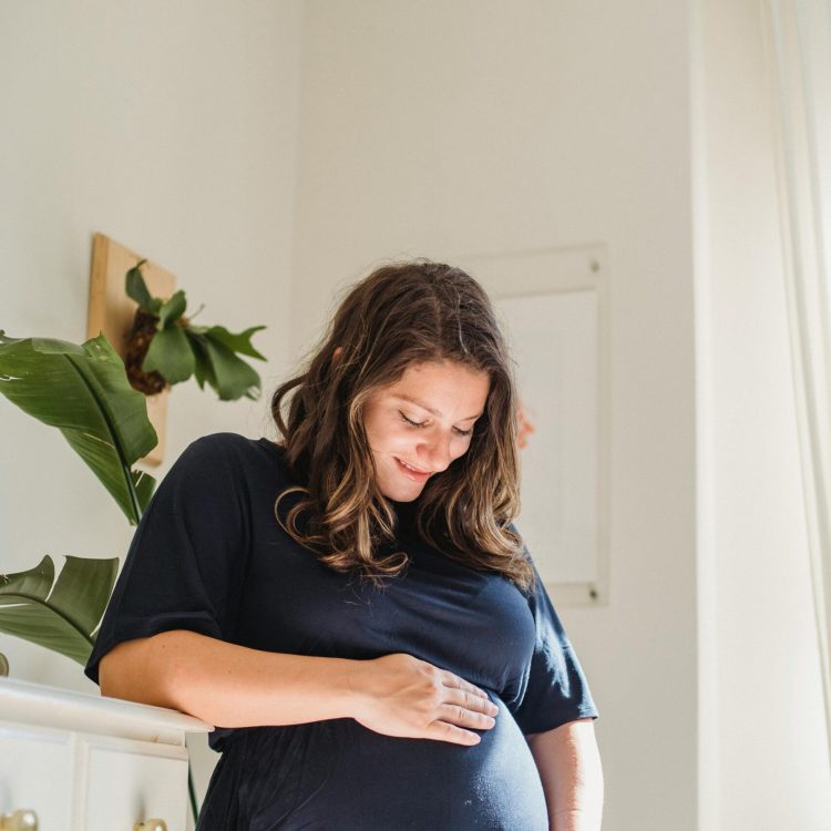 Pregnant woman with brown hair and a blue dress holding belly in her hands, looking at baby bump, and smiling | Unexpected Pregnancy | Mary's Refuge | Martinsburg WV